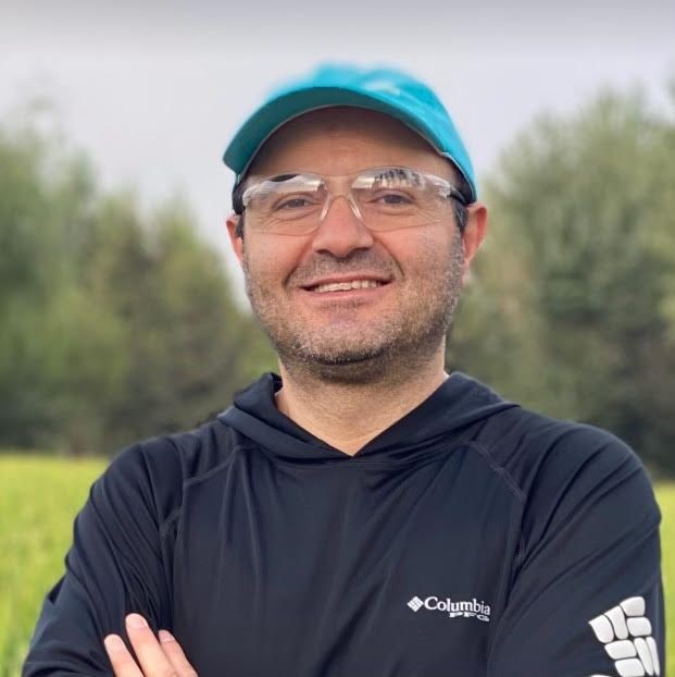 Person wearing a blue cap and transparent glasses, standing outdoors with trees in the background.